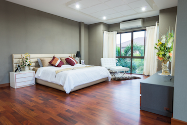 a large room with a hardwood floor, a tall window overlooking trees, a two-person be with a single bedside table, and a padded chair next to the window
